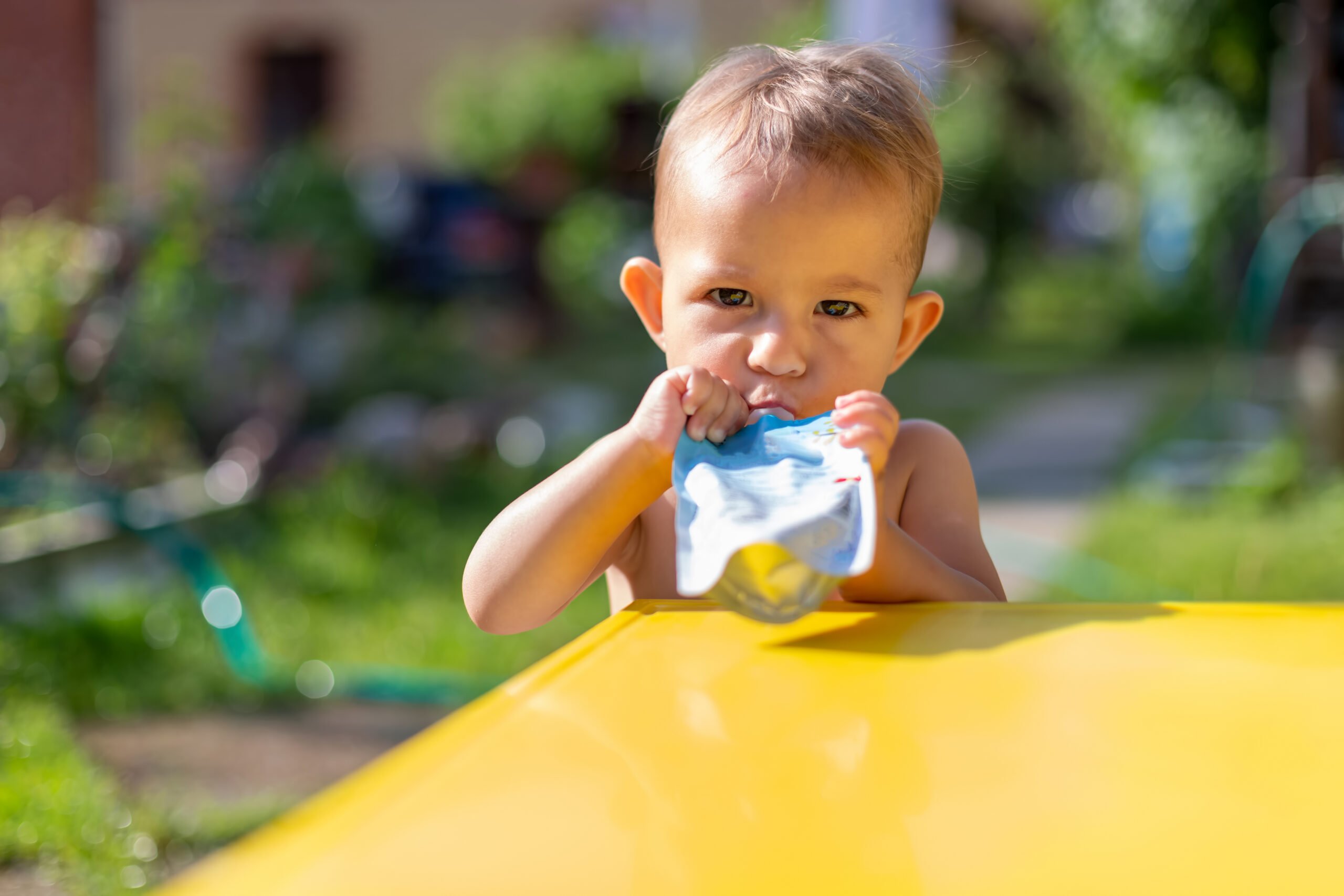 serious baby eating fruit puree in baby food pouch and looking into the camera in front of the yellow table. on the background is a green garden on a sunny day in blur