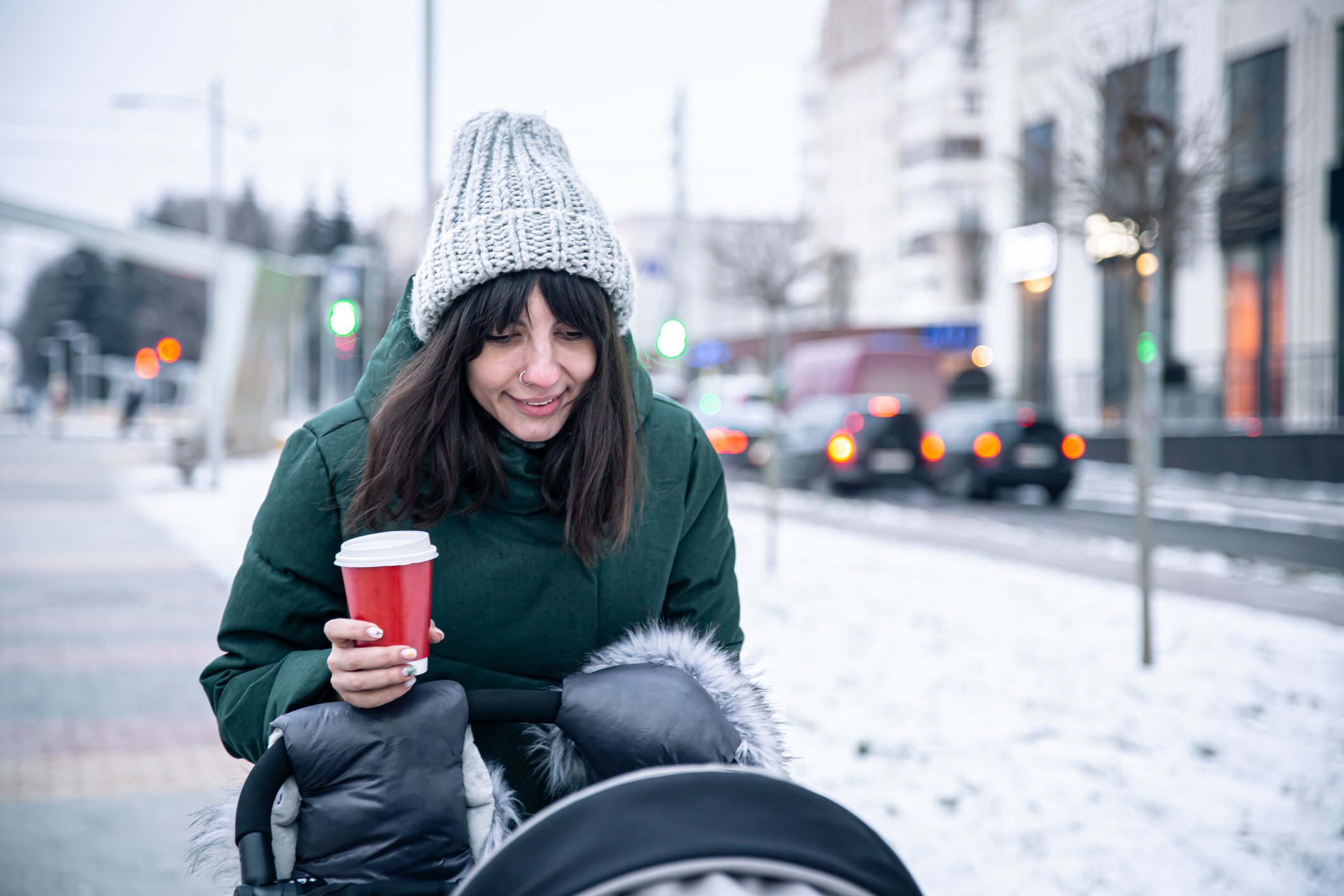 Stylish young mom with a cup of coffee on a walk with a baby carriage in winter.