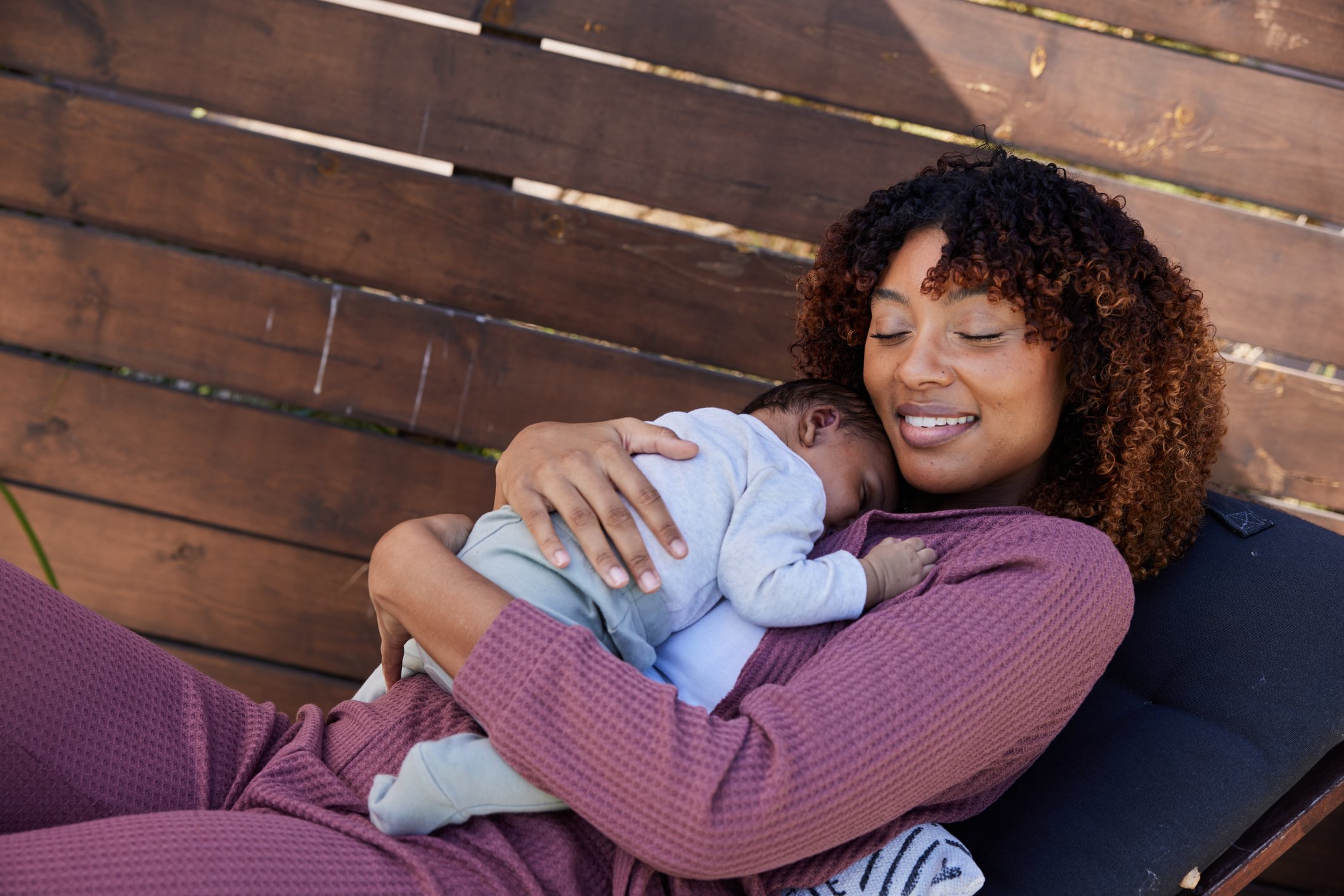 Smiling mother lying with her eyes closed outside on her patio with her sleeping little boy in her arms