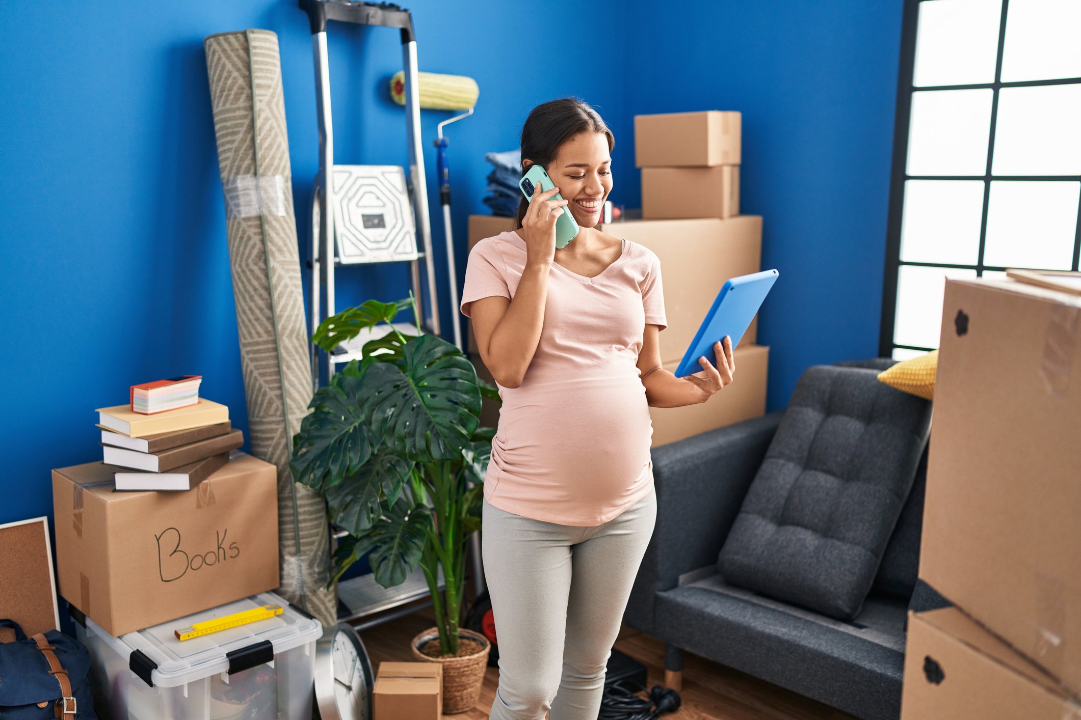 Young latin woman pregnant using touchpad talking on the smartphone at new home