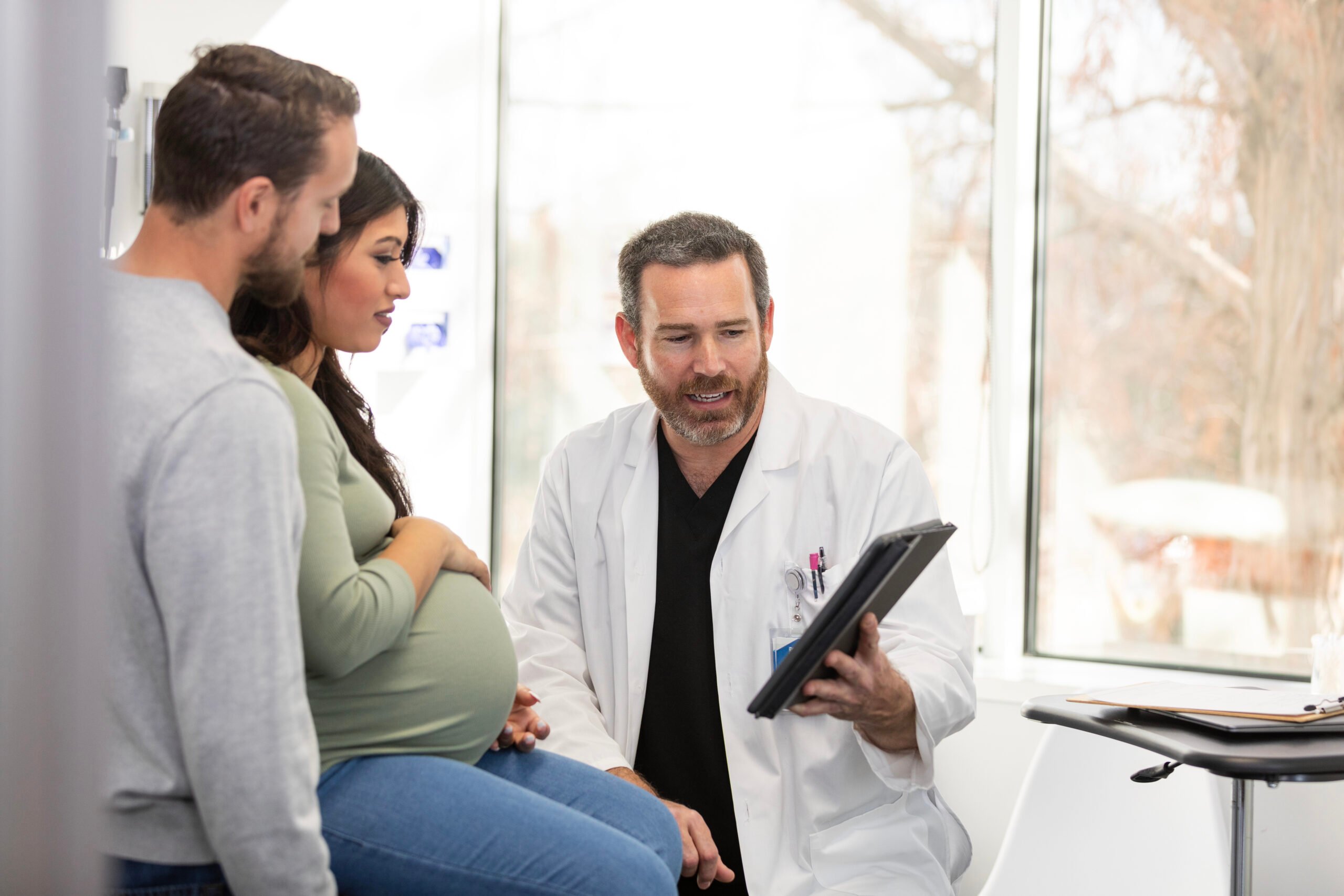 The mature adult male obstetrician uses his digital tablet to show the expectant couple the results of their medical tests.