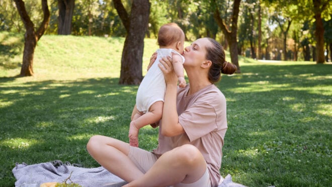 Loving mother kissing her infant child having fun picnic outdoors family bonding during vacation sitting on the grass enjoying healthy snacks and the joyful moments of happy day in park