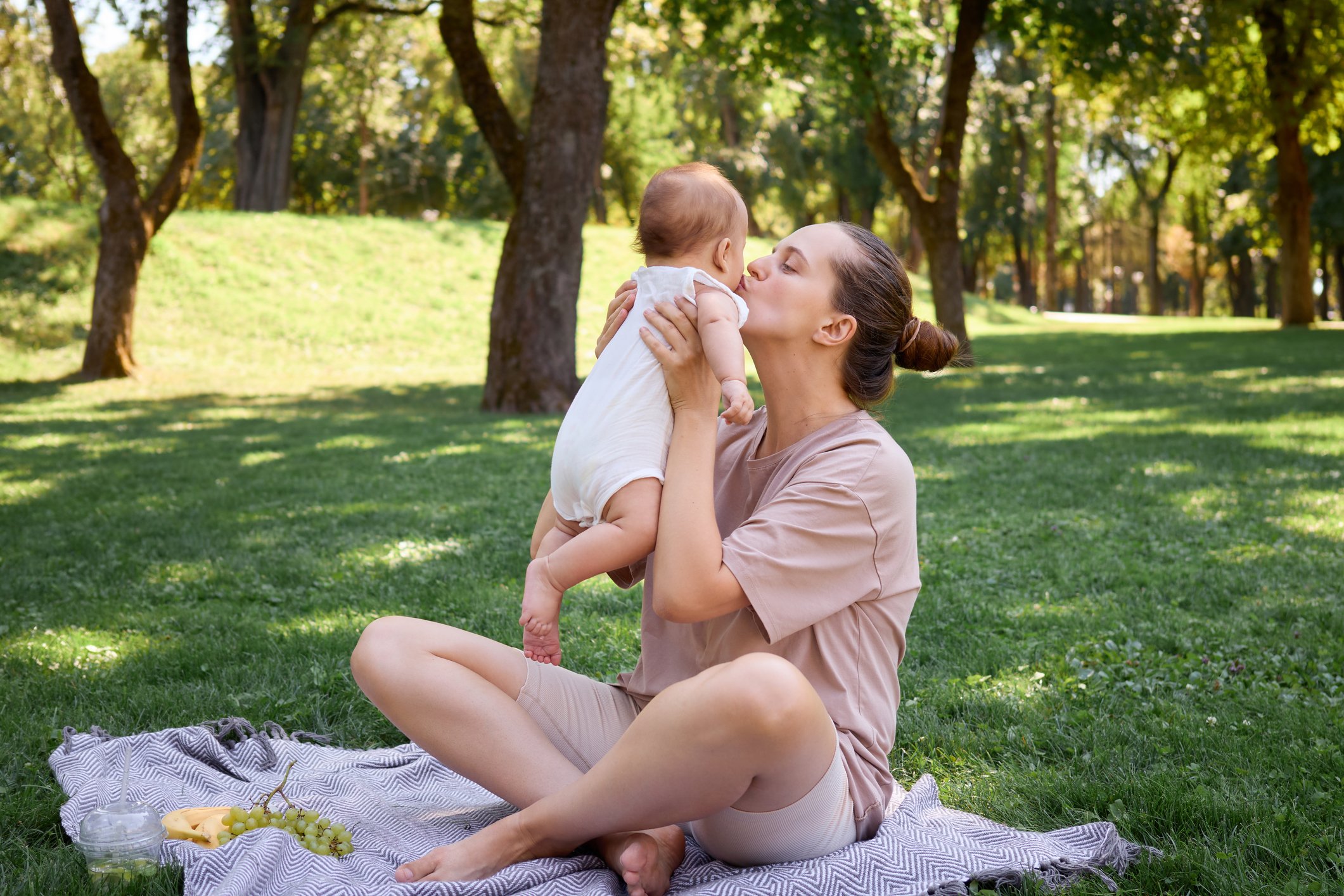 Loving mother kissing her infant child having fun picnic outdoors family bonding during vacation sitting on the grass enjoying healthy snacks and the joyful moments of happy day in park