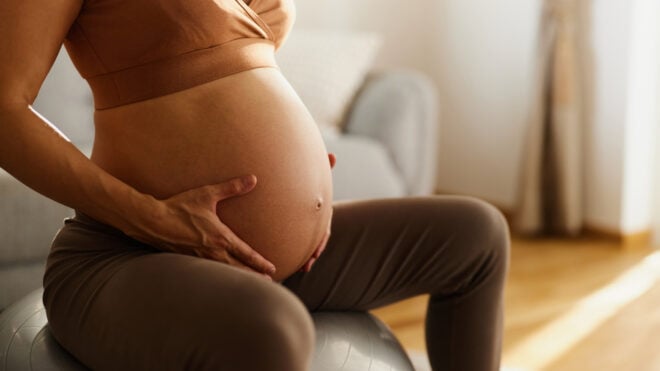Close up of unrecognizable expecting woman holding hands on her stomach while relaxing on a fitness ball in the living room.