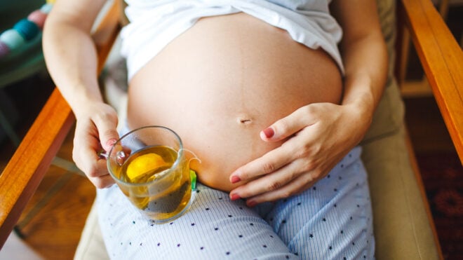A serene shot of an expecting mother resting in a chair, her hand gently holding a glass cup filled with herbal tea.