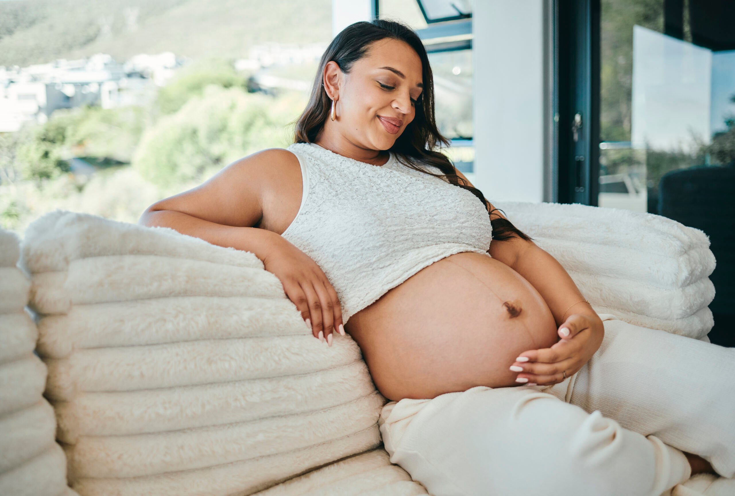 A joyful pregnant woman with long dark hair, sitting on a cozy sofa in a bright living room, gently cradling her belly and smiling warmly.