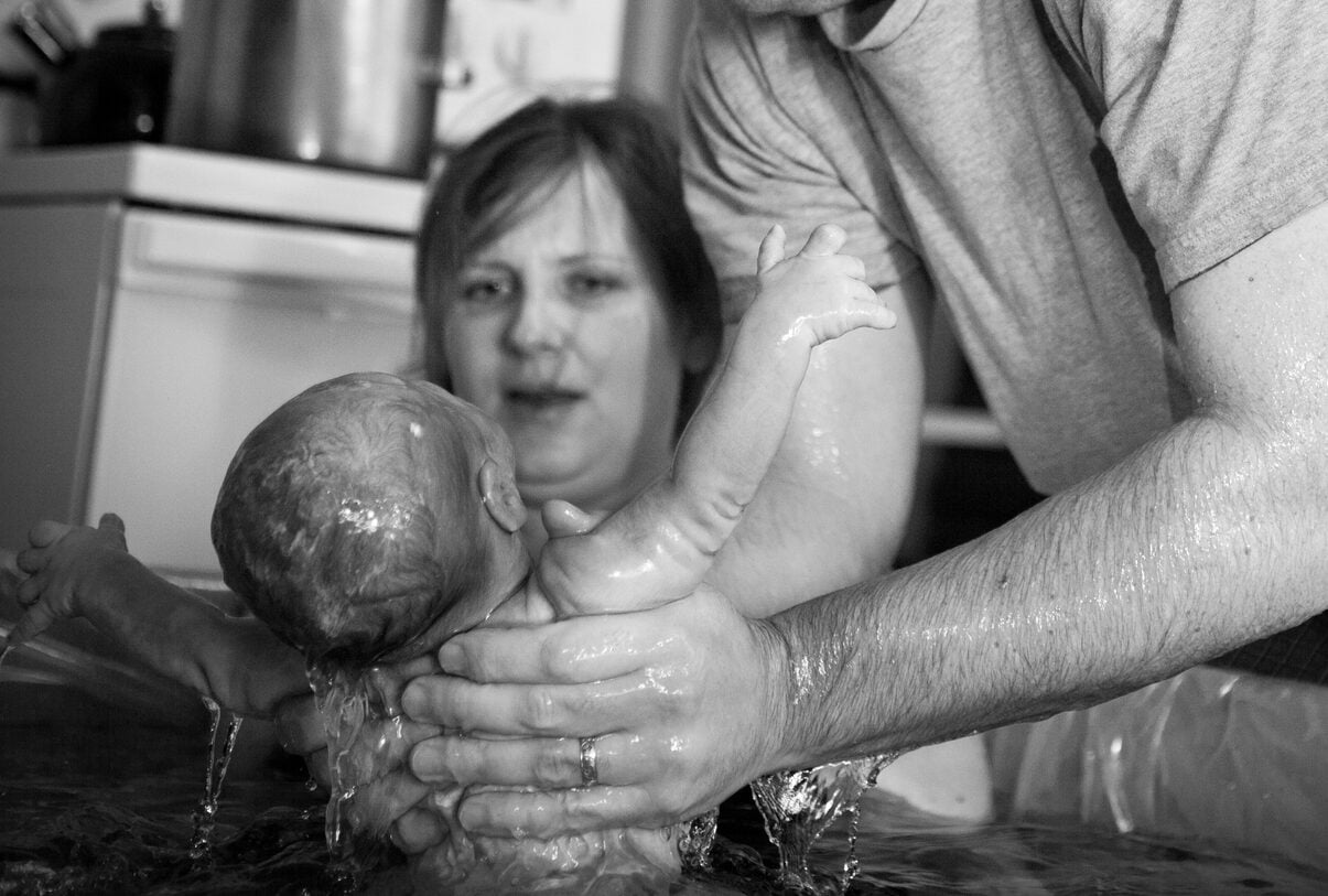Black and white image of a loving husband lifting newborn from water and showing his wife after a home birth.